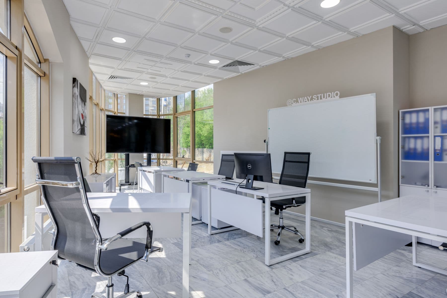 A bright office with white desks, gray chairs, a whiteboard and a wall-mounted screen.